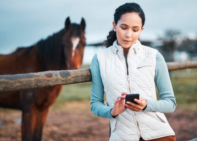 Shot of an attractive woman using her cellphone while posing with a horse in an enclosed pasture on a farm